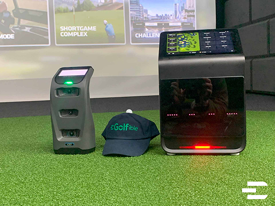 Garmin Approach R50 and Foresight GC3 launch monitors side by side on turf with Golfible hat, in front of golf simulator screen.