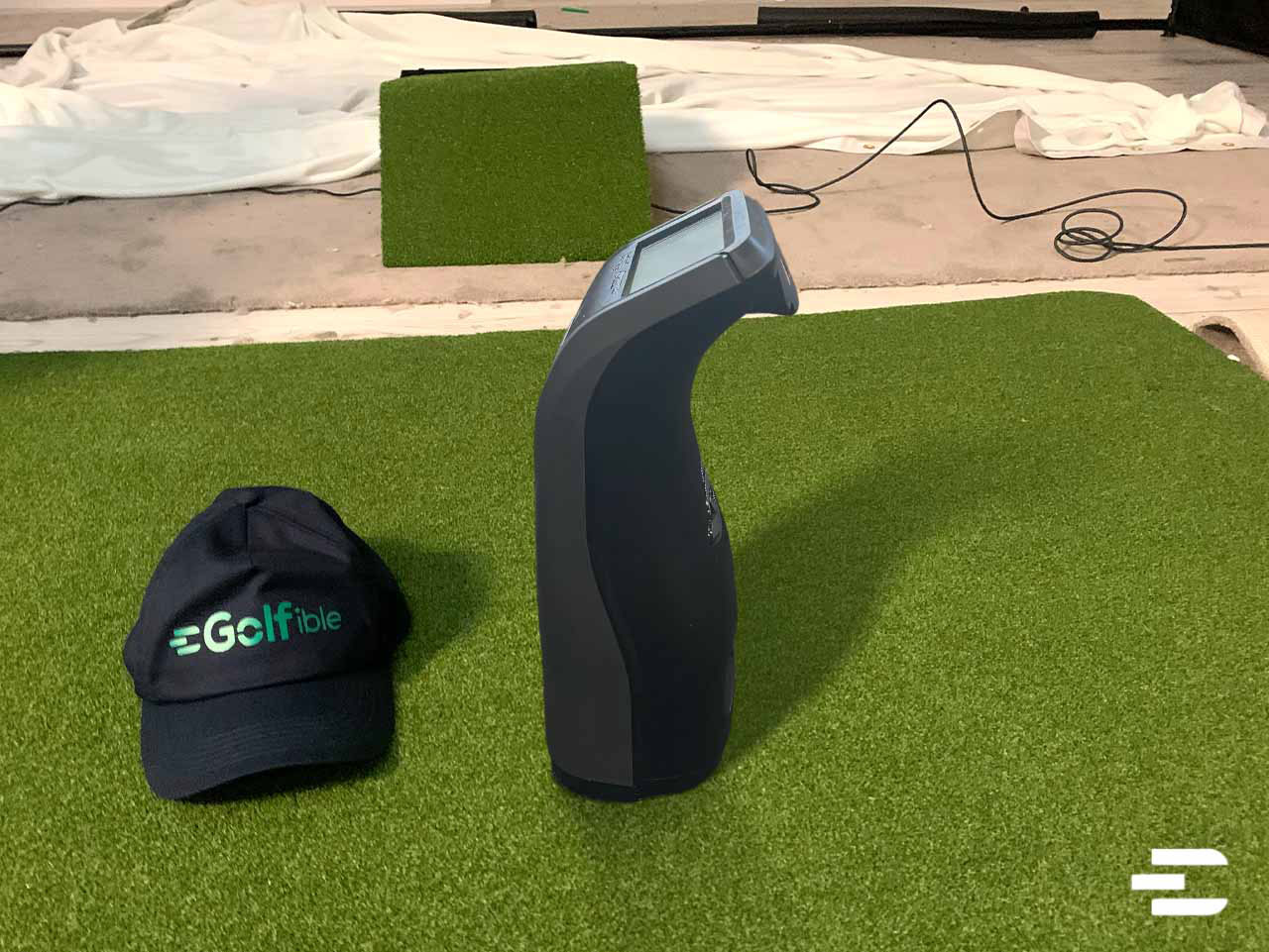 Side view of the Bushnell Launch Pro beside a Golfible cap on a green turf mat indoors.