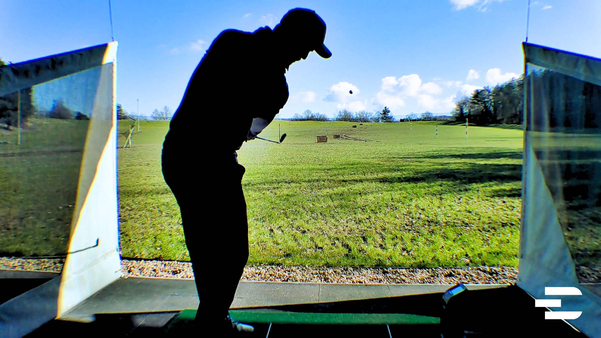 Golfer hitting iron shot outdoors with Bushnell Launch Pro under direct sunlight at driving range