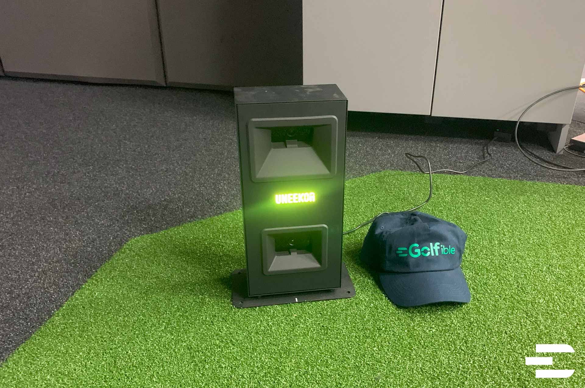Close-up of Uneekor Eye Mini Lite launch monitor glowing green on turf, beside a Golfible cap